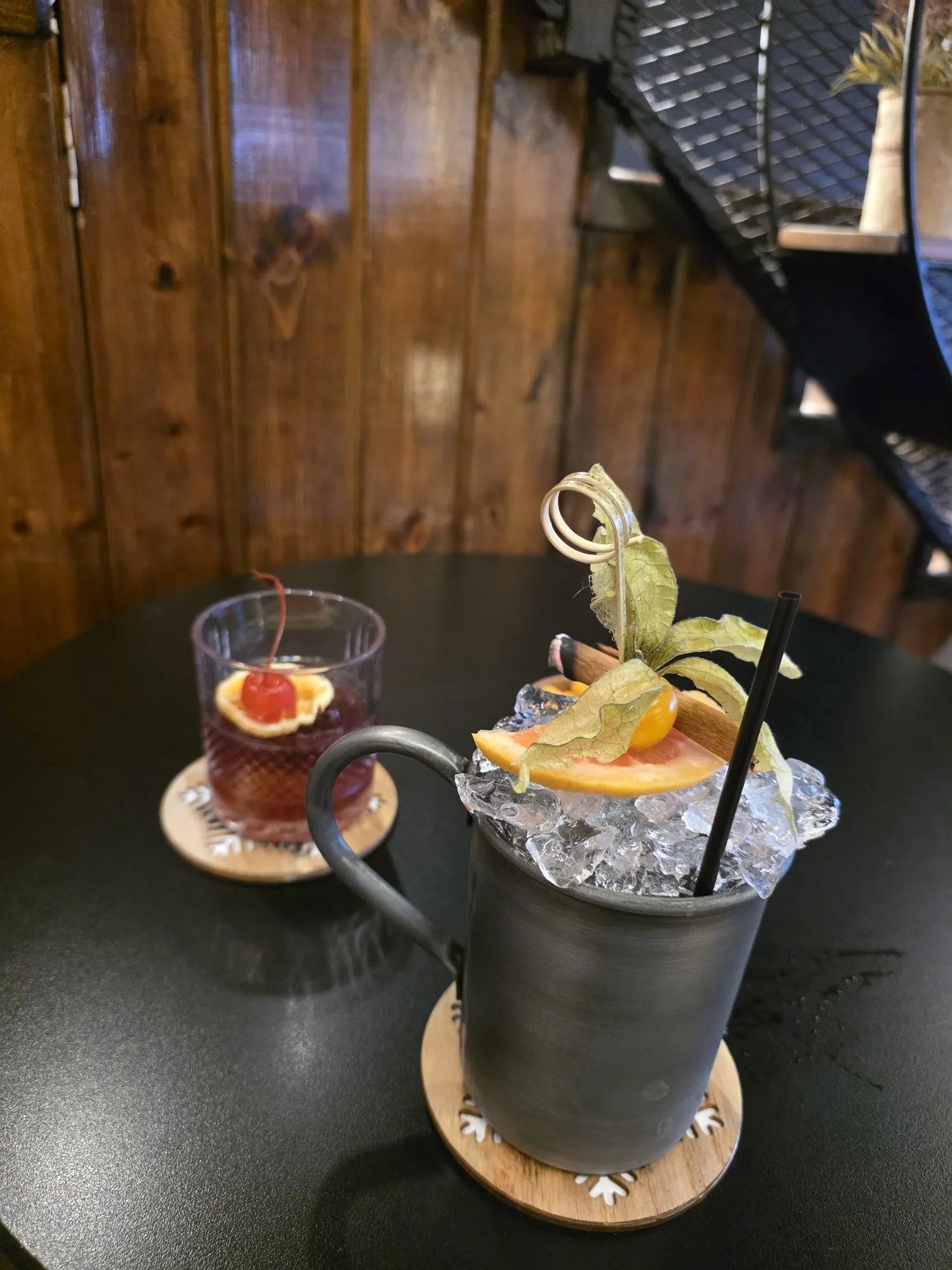 A short drink with orange and a cocktail in a dark metal mug on a black table